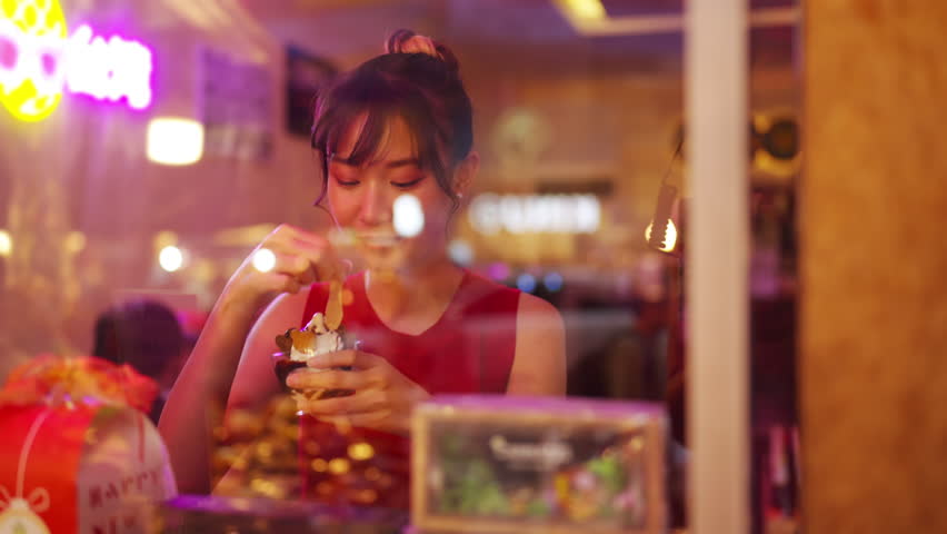 4K Attractive girl sitting by the window at cafe with eating ice cream and looking city street night lights at summer night. Asian woman enjoy and fun outdoor lifestyle in the city on holiday vacation | Shutterstock HD Video #1098482267