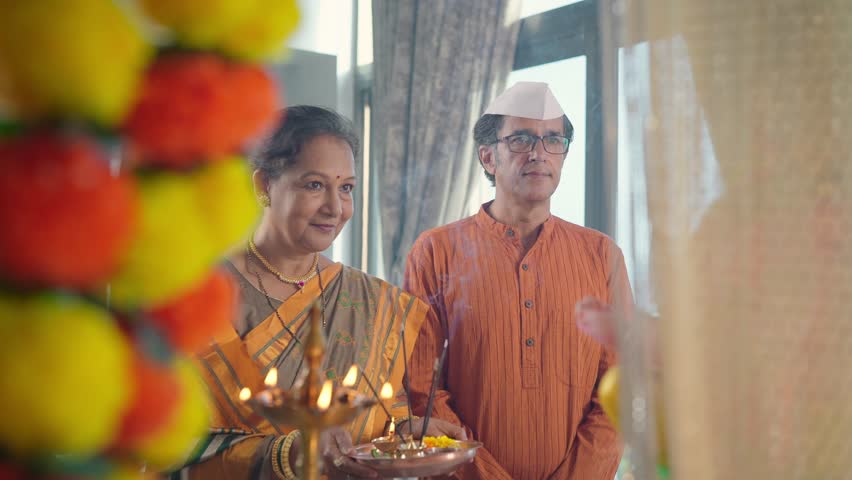 Happy ethnic Hindu Indian old or elderly married couple praying or worshiping the Ganapati idol decorated with marigold flowers during the Ganesha Chaturthi festival or celebration in an indoor house Royalty-Free Stock Footage #1098496311
