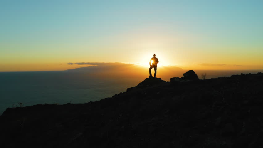 Hiking man in mountains watching sunset and horizon over beautiful ocean landscape. Male hiker silhouette against cloudy colorful sky with sunburst, sun flare.