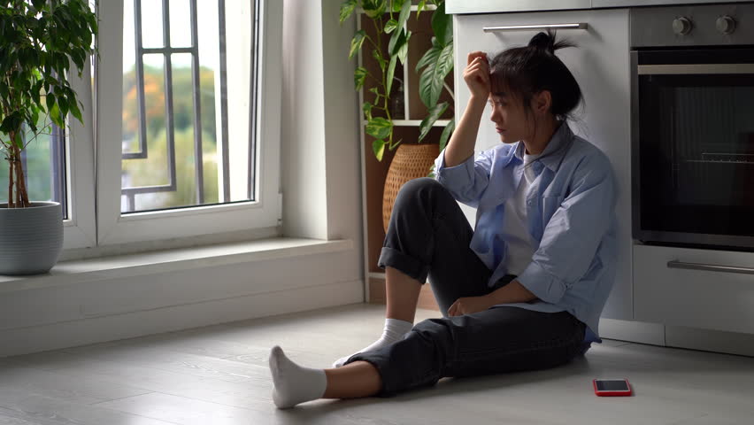 Upset depressed Asian girl suffering from breakup, heartbroken frustrated woman sits on floor in kitchen looking at smartphone waiting for call or message from ex boyfriend. Codependent relationships | Shutterstock HD Video #1098502573