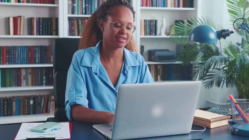 Young optimistic attractive African American woman secretary typing text on laptop keyboard for email messages to partners or clients of company or doing errands for boss sits at table in office | Shutterstock HD Video #1098505609