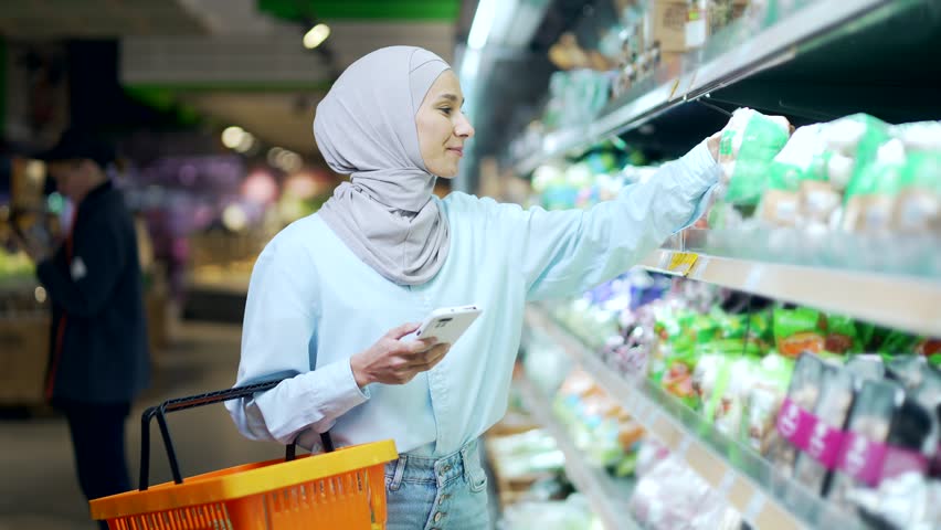 Young muslim woman in hijab Buyer customer checking shopping list using smartphone, purchasing in grocery store in supermarket female shopper pick goods use mobile phone in hypermarket or food market | Shutterstock HD Video #1098506289