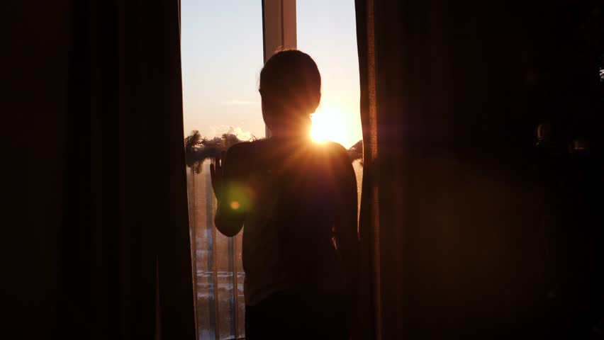 Young girl child go to panoramic window in house in evening, open curtains with hands so sunlight illuminates cozy room at sunset. Warm sun rays shine into camera and glare. Put hand palm on glass | Shutterstock HD Video #1098513627