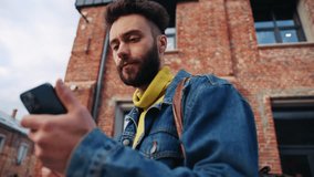 Shooting from below of focused young bearded man holding smartphone in hand, reading message. Portrait of attractive guy with dark hair using mobile phone. Outdoor. Timelapse shooting - Powered by Shutterstock - Get 15% off with code: PIKWIZARD15