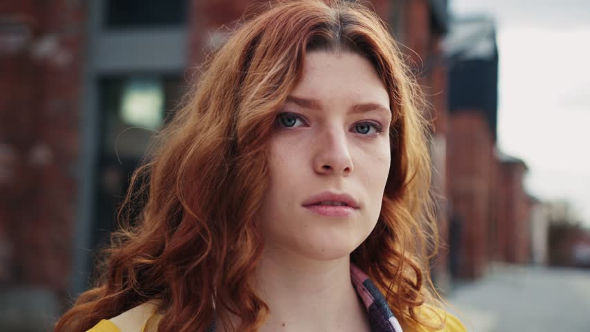 Close-up view of good-looking serious young woman with ginger hair looking at camera. Portrait of calm redhead girl with green eyes on blurred background outdoor. Pensive expression | Shutterstock HD Video #1098517087
