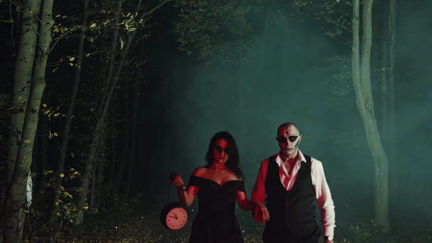 Spooky couple of ghosts man and woman dressed in black classic clothes wandering in the woods at night with a clock stopped. High quality 4k footage