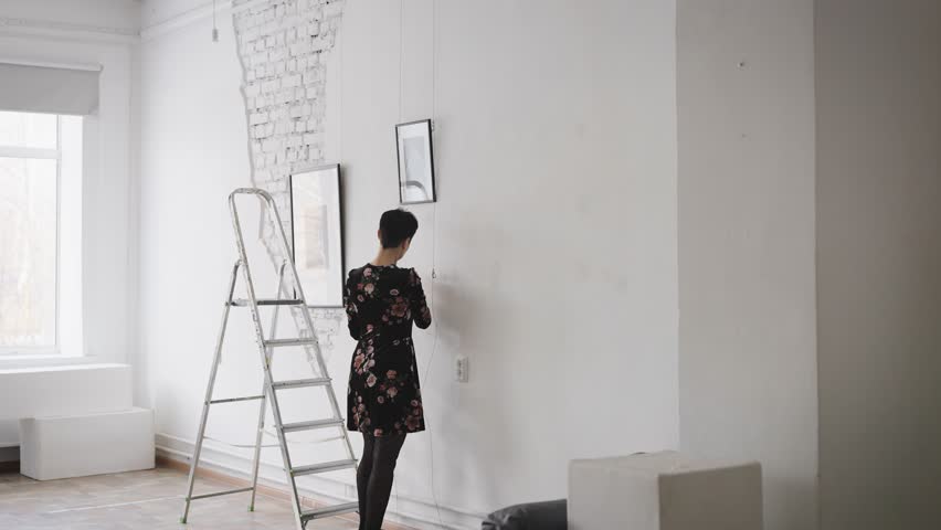 A woman gallery owner hangs new paintings and collages on the wall of the exhibition hall. Gallery with an exhibition of contemporary art.