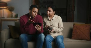 Happy Latin couple talks together about interesting video on smartphone. Woman drinks hot beverage and enjoys spending weekend with boyfriend