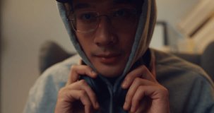 Close-up young Asian man gamer wear gray hoodie and red cap focus on screen put headphone with serious moment ready to competition play video game sit on sofa living room home at night.