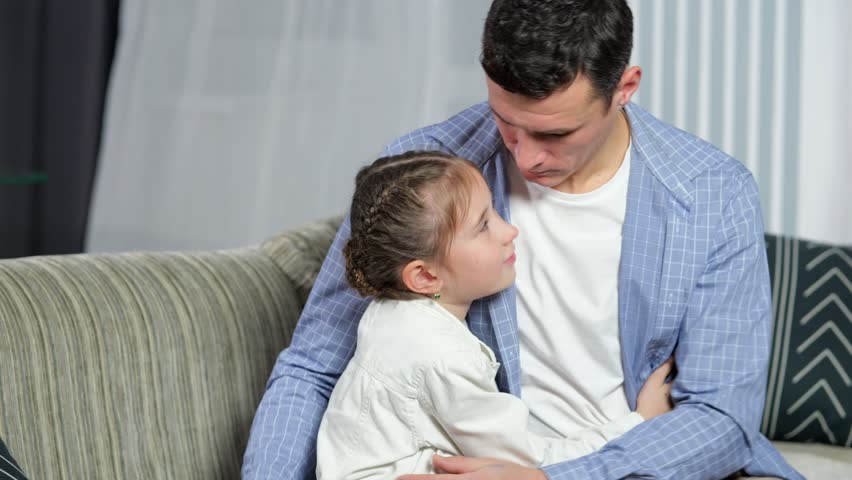 Loving father shows love and embraces precious daughter at home. Lovely junior schoolgirl kisses father on cheek and hugs man tight on sofa in living room Royalty-Free Stock Footage #1098536035