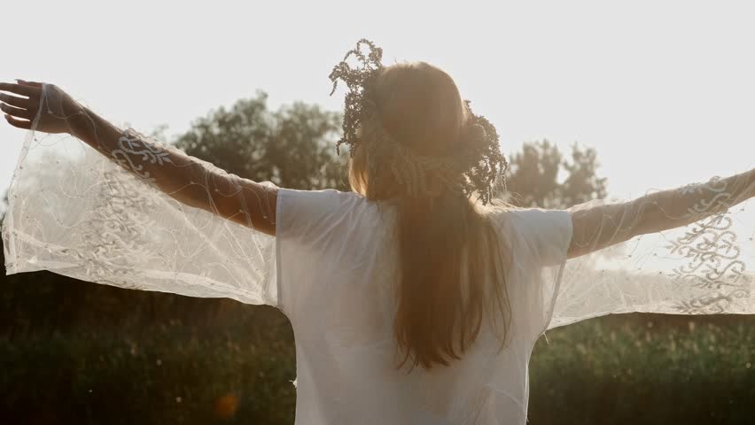 A young woman in a white dress and a wreath of wild flowers raises her hands up in the rays of the bright sun. Back view
