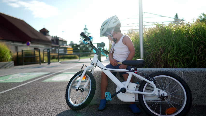 Young boy cyclist wearing helmet resting sitting by bike lane. Child little kid seated by bicycle in sunlight