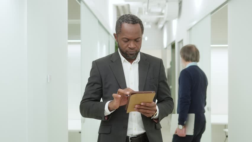 Portrait of successful serious young African American man using tablet walking in office. Handsome focused worker of modern IT company wearing smart style clothes using technologies online. | Shutterstock HD Video #1098542807