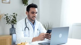 Cheerful ethnic doctor using smartphone while working on laptop at home