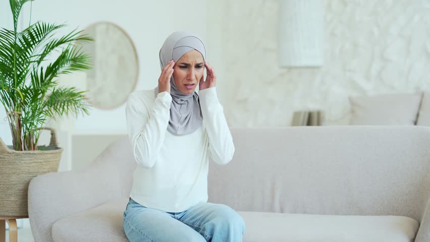 Unhappy young Muslim woman in hijab sitting on couch rubs temple with closing eyes suffering headache painful feelings chronic migraine high or low pressure Stressed upset female feels hurt at home | Shutterstock HD Video #1098561493
