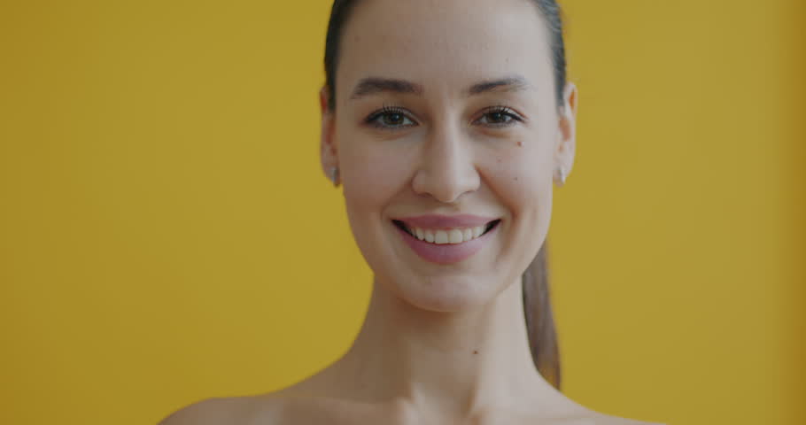 Slow motion portrait of young woman having fun with cucumber slices implying skin care with natural mask smiling looking at camera on yellow background