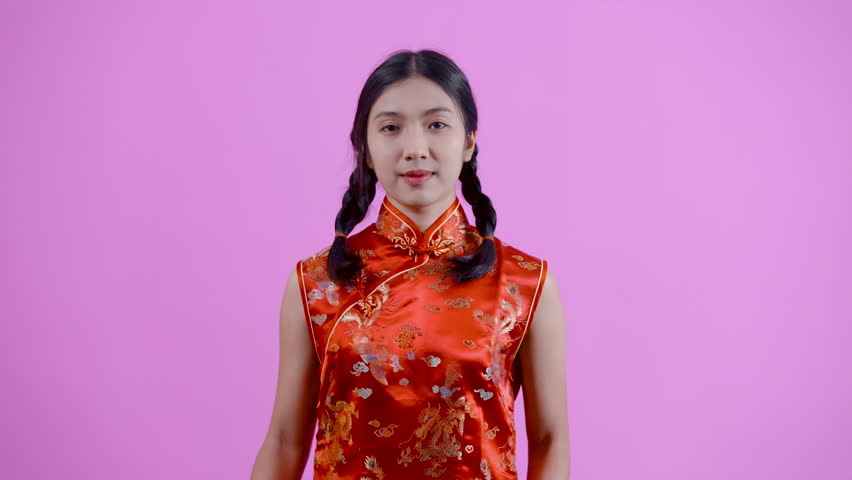 4K, Asian woman of Chinese descent with pigtails wearing red chinese new year cheongsam Happily standing and dancing with two thumbs up. Smiling, having fun, alone, studio isolated on pink background