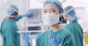 Video portrait of asian female surgeon in cap, gloves and face mask in operating theatre, copy space. Hospital, medical and healthcare services.