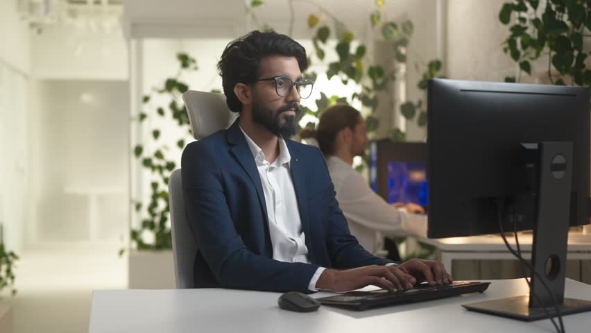 Portrait of young professional Indian IT company worker typing on keyboard using computer in modern office. Focused bearded happy man developer coding wearing suit looking at camera smiling. | Shutterstock HD Video #1098580793