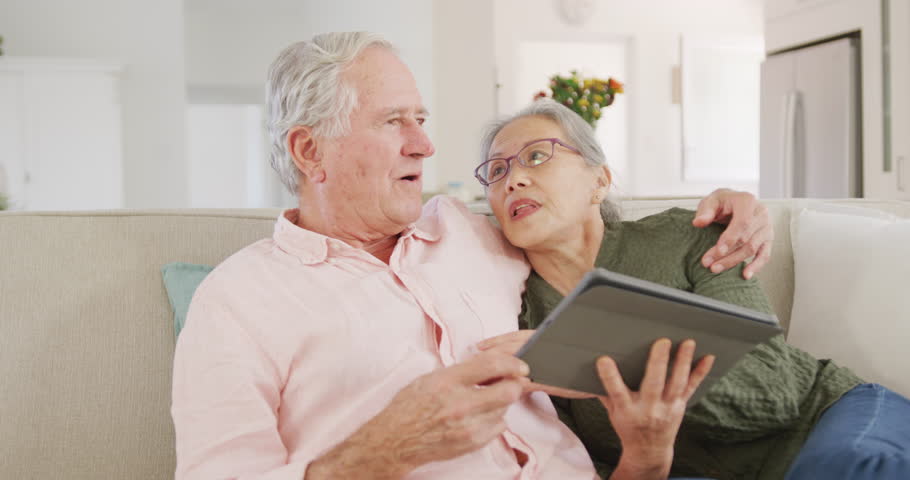 Happy diverse senior couple using tablet and sitting on couch. Active retirement and lifestyle concept.