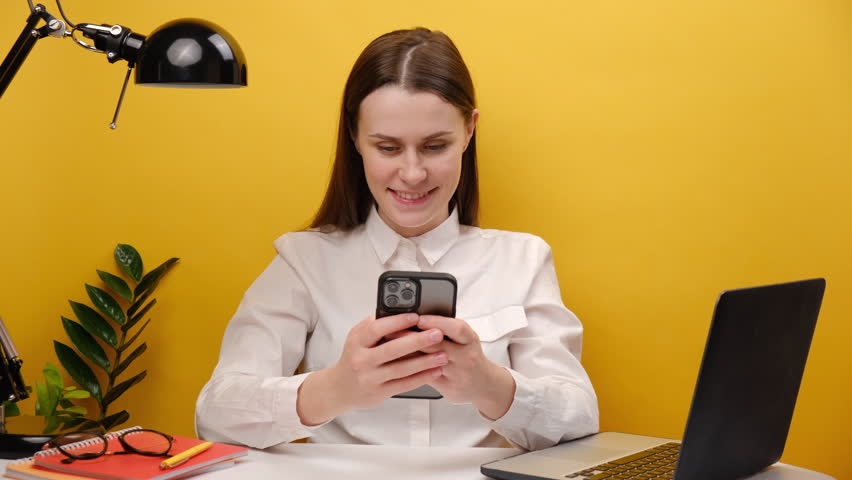Portrait of beautiful successful employee young woman sit work at white office desk with laptop holding mobile cell phone browsing surfing internet, posing over yellow color background wall in studio | Shutterstock HD Video #1098587621