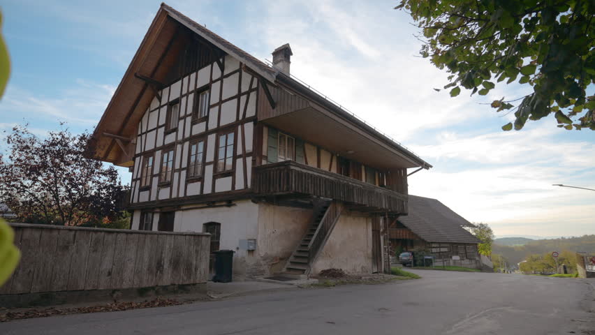 Mother holds hand of toddler boy walking on road past vintage two-story village house. Woman and toddler enjoy sunset in Swiss countryside backside view slow motion