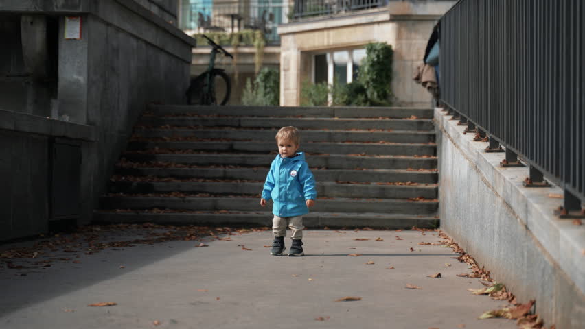 Cute blond toddler walks back to stairs at family walk around city. Little boy in blue jacket explores new places during autumn trip in Switzerland