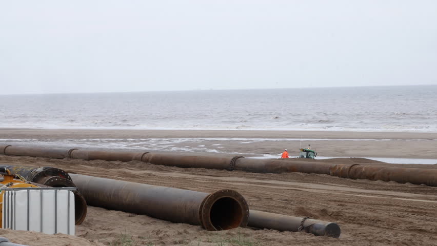 Coastal pipeline contructions. Pipes are laid out on the beach with construction workers digging a trench near to the sea in the UK