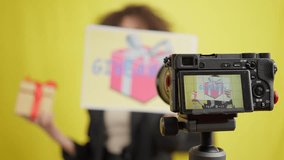 Close-up video camera screen with blogger advertising gifting and blurred young woman talking at yellow background. Positive charming brunette Caucasian vlogger filming surprise offer