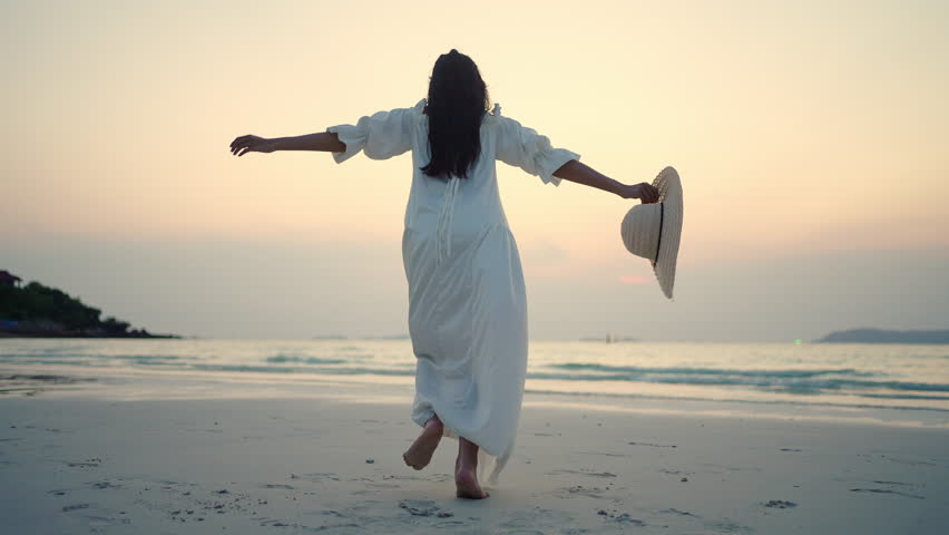 Young Asian woman in white dress running to the ocean and jumping during travel tropical island beach on summer holiday vacation at sunset. Attractive girl enjoy outdoor lifestyle walking at seaside.