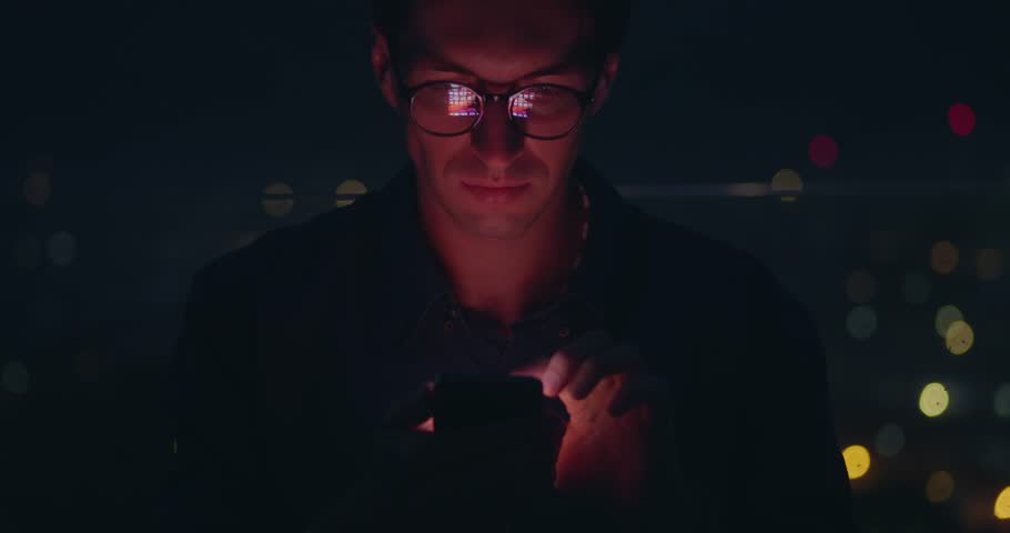 Portrait of young man in glasses scroll internet pages social network on smartphone, on background of night city. Smartphone screen reflected on man's face and glasses. Concept of smartphone addiction