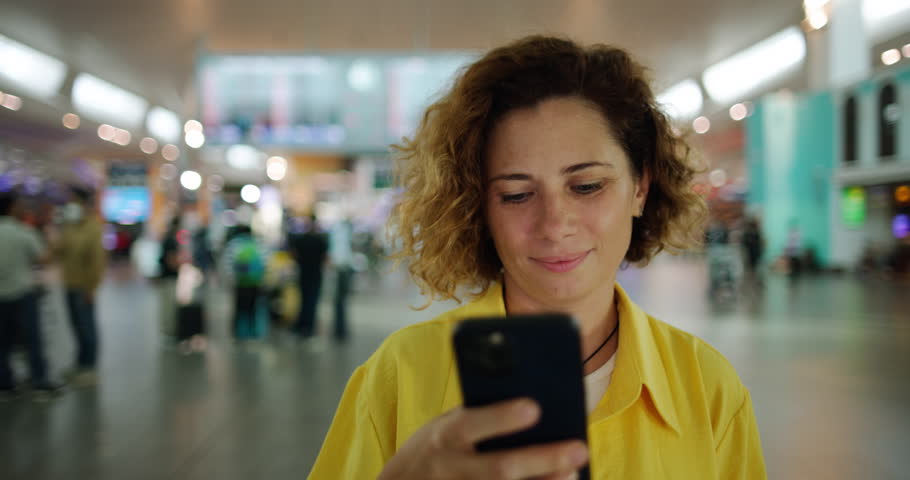 Caucasian woman with brown curls looks at the phone, standing in the middle of the airport. A woman at the airport issues tickets for a flight online. A woman buys plane tickets using her phone. Royalty-Free Stock Footage #1098600149