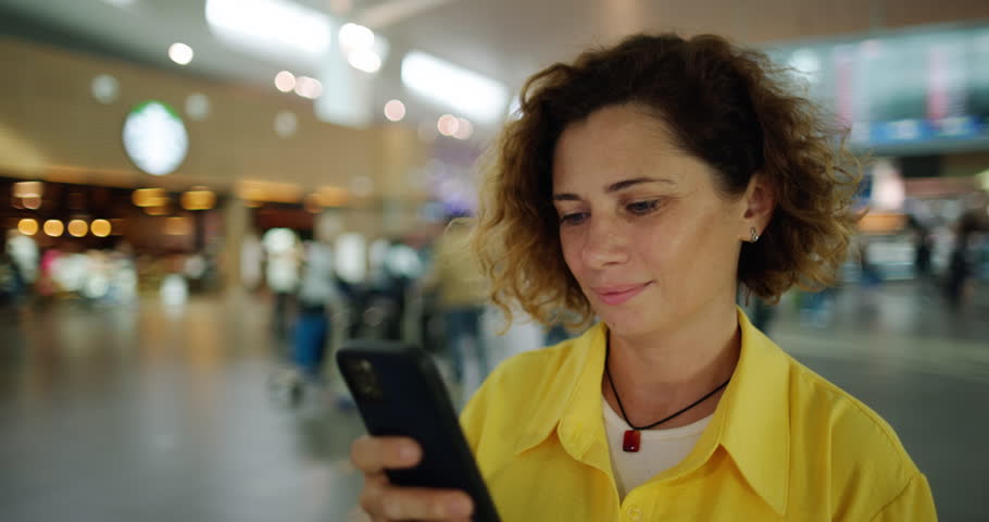 Caucasian woman with brown curls looks at the phone, standing in the middle of the airport. A woman at the airport issues tickets for a flight online. A woman buys plane tickets using her phone.