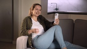 
Happy young woman sit on sofa at home holding phone looking at screen waving hand video calling distance friend online by mobile chat app using smartphone videochat application.