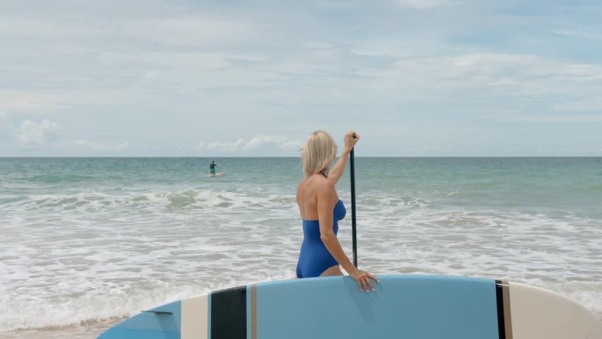Middle age woman in swimsuit stands on the beach, holds a sub board and a paddle, next to the ocean 
