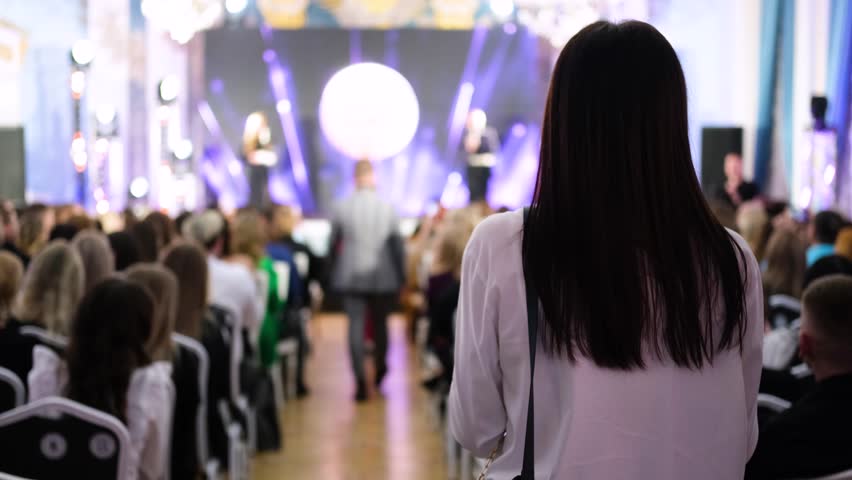 Guests in evening attire sit in a spotlighted lobby and look at the stage. The girl stands with her back to the camera and watches the event Royalty-Free Stock Footage #1098617143