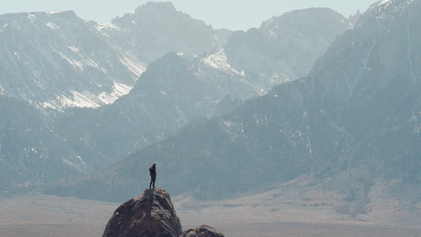 Young adventurous hiker standing on rocky summit looking at massive mountains