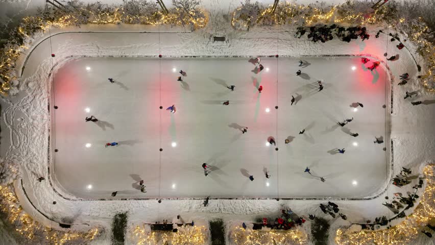 Rising overhead drone shot of people skating around in a circle on festive outdoor skating rink at night