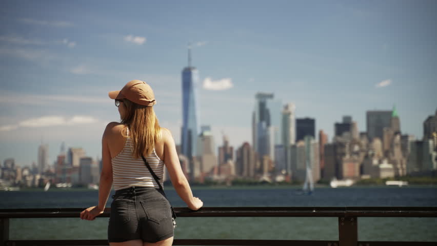 A video showing the back view of a woman standing in place and resting her hands on the black steel railing, looking at the New York city skyline across the river during daytime