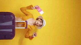 Vertical video. Vacation excitement. Dancing woman. Travel inspiration. Happy lady moving in rhythm posing suitcase and boarding documents on yellow background.