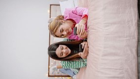 Vertical video of two adorable positive sincere teenage girls, best friends sharing happy time together while visiting each other, sharing social content, gossiping, reading funny news on smartphone