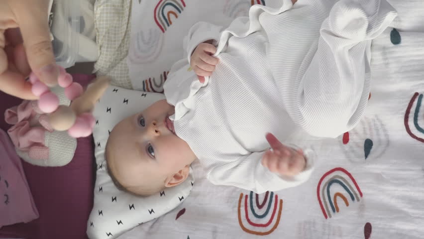 Vertical video. Adorable baby girl playing with rattle toy while lying on back on changing table. Development games for infant with mother