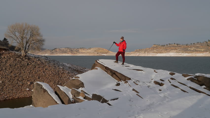 senior, male hiker with trekking poles is standing on a rocky shore of a mountain lake in front of a camera and talking to a wireless microphone - vlogging concept