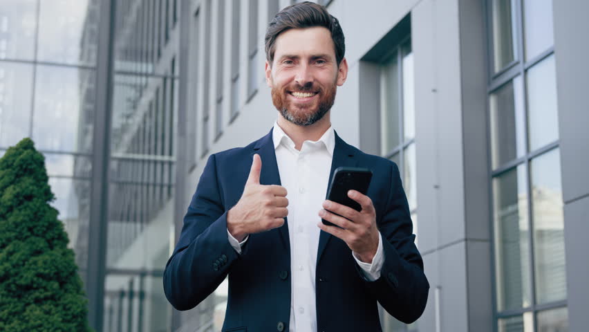 Smiling man holding phone checking email message reading good news happy businessman using mobile application on smartphone looking at camera showing thumb up gesture approval excellent result sign Royalty-Free Stock Footage #1098651213