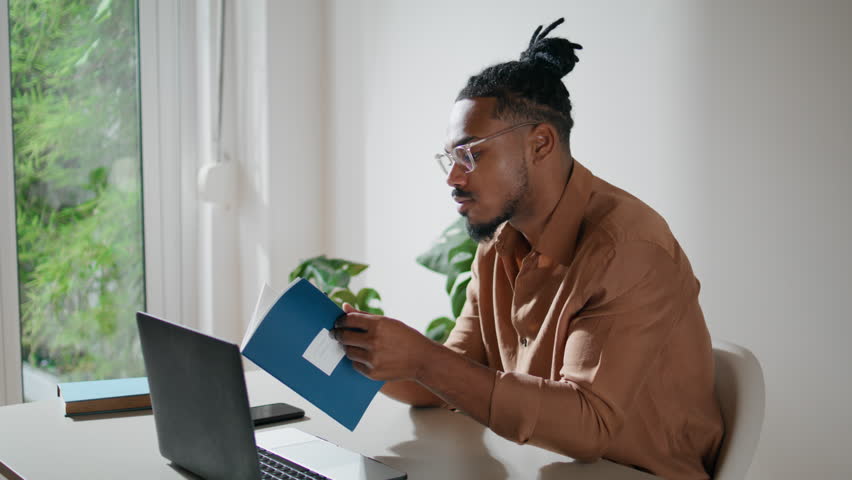 Modern student learning remotely closeup. Stylish guy making notes at home. Focused freelancer watching webinar writing in notebook alone. African american man using laptop computer in light space  Royalty-Free Stock Footage #1098655517