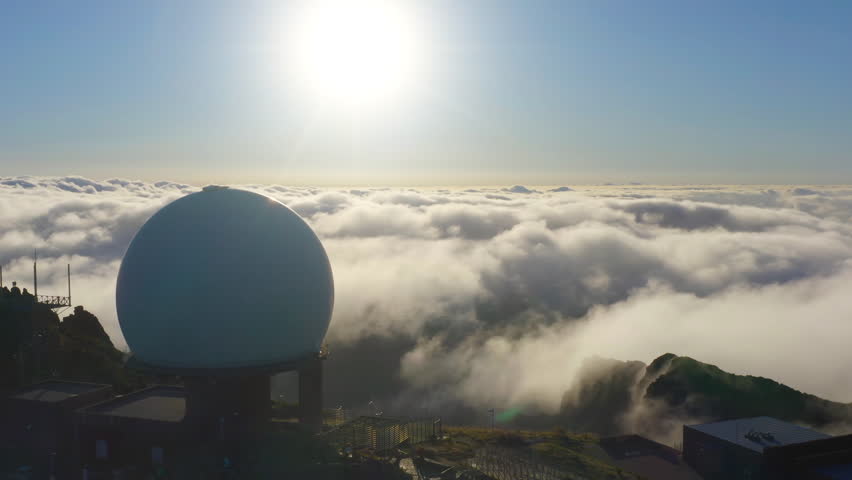 Meteorological weather radar station with a large white sphere on top of a mountain over a clouds at sunset. Aerial view