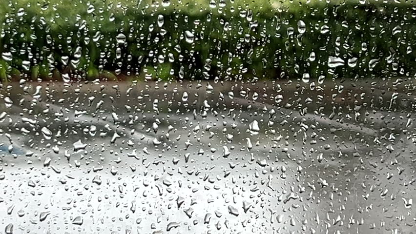 4K HD video close up of water, rain drops on window running down the glass. Rain out of focus in background on parking lot with hedge border.