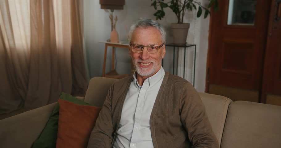Mature man in glasses sits at home and demonstrates perfect smile. Old pensioner with excited expression enjoys spending life in retirement | Shutterstock HD Video #1098663155