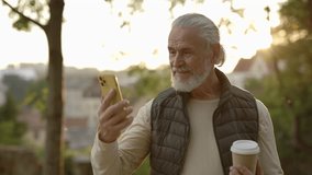 Sporty mature man chatting with friends on online video call on smartphone standing in the part. Elderly male using smartphone outside. Video conference. People and technology concept