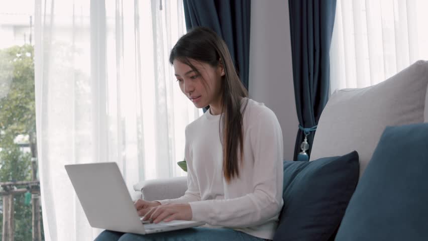 Happy beautiful young asian woman smile sitting on sofa and holding mobile relaxing at home. Beautiful girl happily listening to music on a sofa inside a home office using a wireless headphone. | Shutterstock HD Video #1098666325